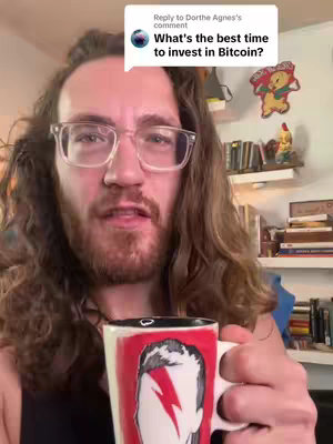 A man with long, wavy brown hair and transparent glasses speaks directly to the camera in a room with books and collectibles. He is holding a mug showing a red face with a lightning bolt and is wearing a black shirt. The background includes shelves with books, and a comment bubble above his head reveals an inquiry about the best time to invest in Bitcoin.