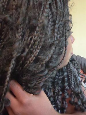 Close-up shot showing the side profile of a person's head and upper body. Their head and shoulder are visible. The hair is styled in dark, grey-toned braids and curly sections. The person's skin is visible, and they are wearing a dark-colored top.