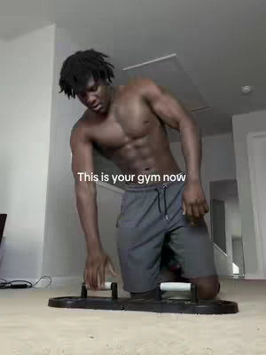 A shirtless Black man with a muscular build is shown from the waist up, kneeling on the floor. In front of him is a push-up board with handle attachments. The background is a lightly colored room with visible stairs and a door. The text overlay reads, "This is your gym now."