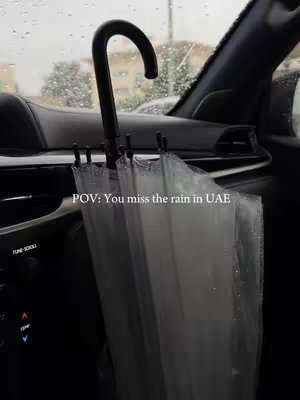 An image captured from inside a car during a rainy day. The focus is on a black umbrella positioned in the car's interior, slightly open and positioned upwards. Raindrops are visible on the car window and in the background, suggesting a view of a wet cityscape. The composition is dark and moody, with muted colors reflecting the cloudy weather.