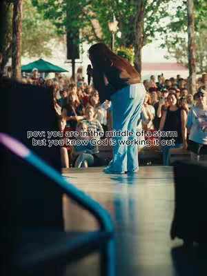 A woman with brown hair stands on a stage, holding a microphone and looking toward the crowd. She's wearing a brown jacket, blue jeans, and is positioned in front of a large audience. Text overlayed across the image suggests the message is about faith and encouragement during difficult times. The scene is filled with warm light and has people in the background.