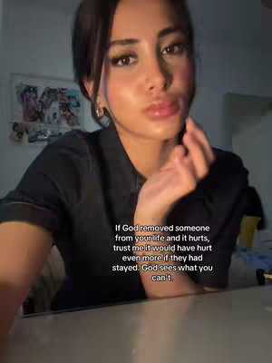 The image shows a close-up of a woman with dark hair and makeup. She is looking directly at the camera, with a slightly empathetic look, almost like she is giving advice. The background is dark and out of focus, with some artwork visible behind her. The lighting is soft, and the overall composition is intimate.