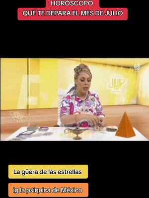 The image is a still from a video, with a woman in the center seated at a desk, presumably indoors. She wears a colorful floral top and has her hair styled up. The background is a muted yellow with some subtle design. On the desk are several objects including a small lamp, cards, and an orange geometric shape. Text overlays at the top and bottom of the image provide context.