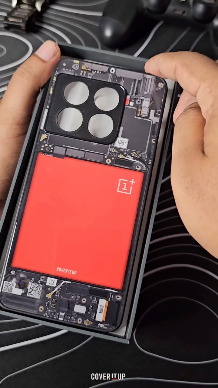 A hand holding a clear phone case that displays the internal components of a phone, set against a blurry background of a dark surface. The case reveals a red rectangular shape, suggesting a battery, alongside other internal structures and the OnePlus logo.