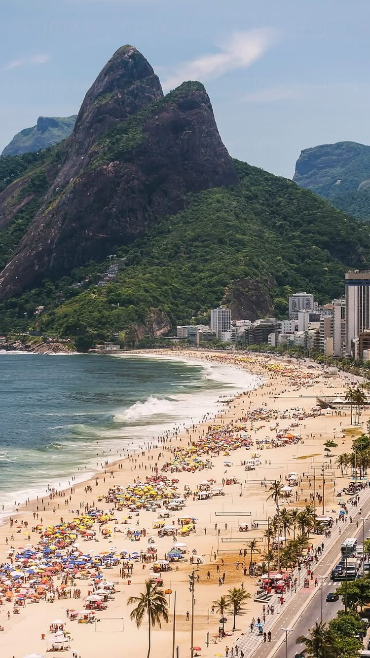 An aerial view of a crowded beach in Rio de Janeiro. The beach is filled with people, colorful umbrellas, and palm trees. Behind the beach, there's a long road with cars and high-rise buildings on the right and a lush green mountain in the background.