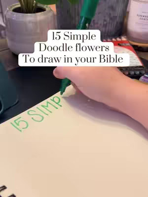 The cover image shows a close-up of a hand holding a green pen, writing on a white sheet of paper. The paper is on a table, and the background includes a blurred view of some potted plants and other items. The writing on the paper says "15 Simple Doodle flowers To draw in your Bible."