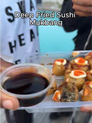 The image shows a person's hand holding a clear plastic container of deep-fried sushi rolls. The sushi pieces are golden brown with a topping of what appears to be white sauce and orange roe. In the foreground, a small, clear container of dark sauce is held as well. The background is blurred and includes parts of a white shirt with black text and a light blue surface.
