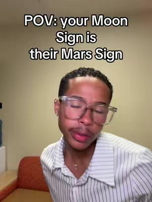 A man with clear-framed glasses is centered in the frame, looking slightly off-camera. The background is a solid, neutral beige wall. Text overlayed at the top reads, "POV: your Moon Sign is their Mars Sign."