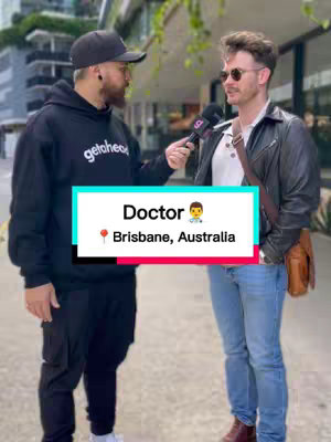 Two men are standing outside on a paved sidewalk. One on the left is wearing a black hoodie and ball cap and holding a microphone, while the second man, to the right, is wearing a black leather jacket and jeans. A white and blue rectangular overlay sits centered at the bottom of the frame.