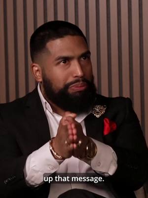 A man in a black suit and white shirt, with a gold watch and bracelet, holds his hands together in front of his chest in a praying gesture. He has a well-groomed beard, and a pin on his suit. Background is a series of vertical white and beige stripes.
