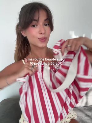 A woman with light skin and brown hair, tied back in a ponytail, is the central focus of the image. She is wearing light makeup, including blush, and appears to be smiling and speaking. She is holding a red and white striped garment, possibly a top or a bag, up towards the camera. The background is a soft, possibly indoor setting, with a glimpse of a patterned yellow top worn by the woman.