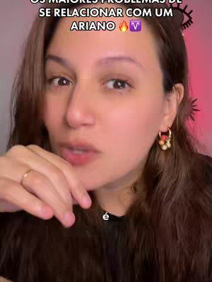 A woman with long brown hair and light makeup is centered in a close-up shot. She is wearing a dark top and gold hoop earrings adorned with colorful beads. A thin gold ring is visible on her finger. The background has a warm pinkish hue. Text overlays are present at the top of the frame.