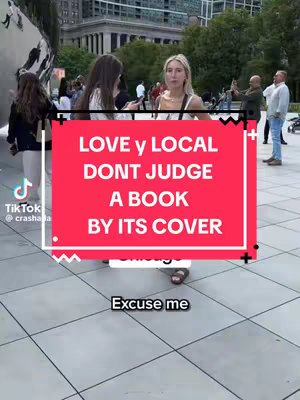 A woman in a white top stands in front of a large, reflective metal sculpture. Overlaying the image is a large red banner with the words "LOVE y LOCAL DON'T JUDGE A BOOK BY ITS COVER". White text below reads “Excuse me”. The background includes a crowd of people engaged in various activities.