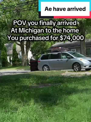 A light-colored minivan is parked on a residential street. A dark green trash can is on the curb beside the car. The background includes green trees, grass, and a house.