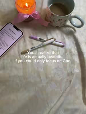 Close-up shot of a white surface with various items arranged on it. The composition prominently features a lit candle in a short glass holder wrapped with a pink ribbon, a coffee cup decorated with floral patterns, and two markers placed diagonally. A smartphone displaying text is positioned to the left, and the overlayed text stating "You'll realize that life is actually beautiful, if you could only focus on God." is centered.