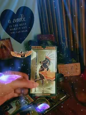The image shows a setup for a tarot reading. A hand is visible, reaching towards tarot cards. In the background, there's a draped curtain, a heart-shaped image with text, and various decorative elements. The lighting is soft and mystical.