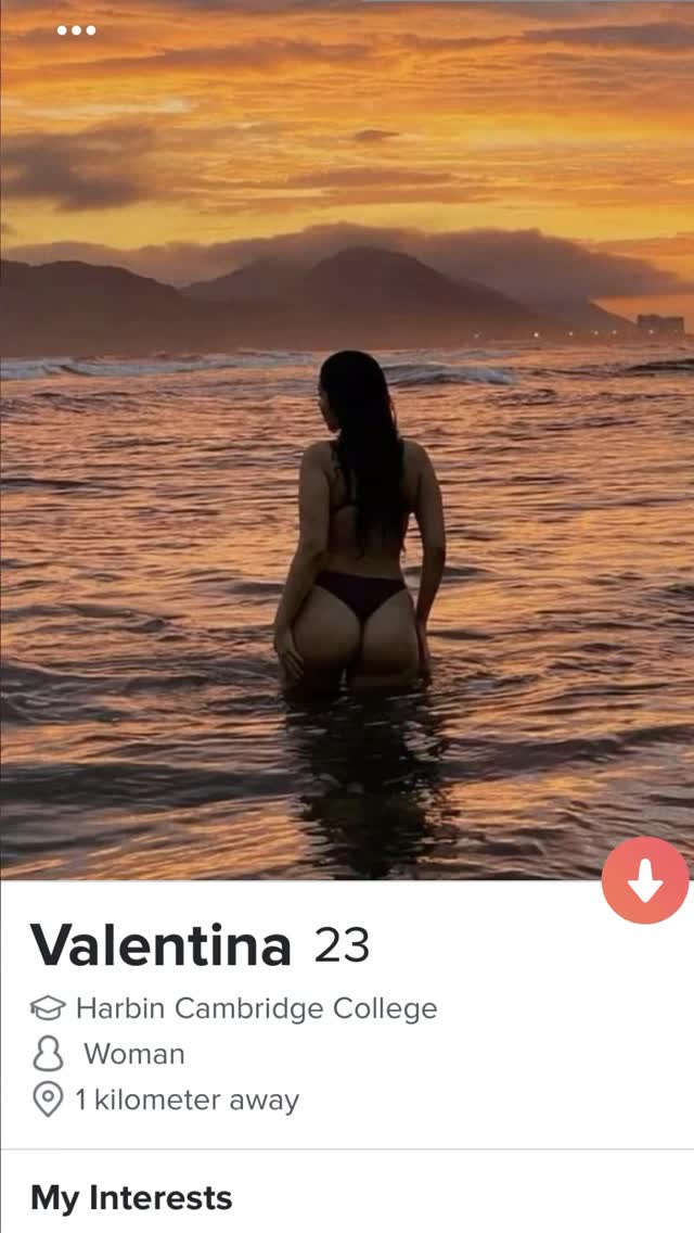 A woman stands in the ocean, viewed from the back. The water reflects the warm colors of the setting sun and some mountains are also visible. The photo has a dating app interface.