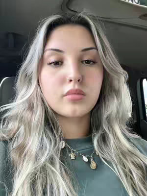 A young woman with long wavy blonde hair is seen in a close-up shot. She is wearing a green top and a necklace. The background suggests she is in a car.