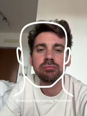 A man with short, dark hair and a beard and mustache is centered in the frame, wearing a white t-shirt. A white outline, in the shape of a face appears over the man’s face, acting as a guide. The hook text overlay reads, "show your face and tap the screen". The background is slightly out of focus, suggesting an indoor setting.