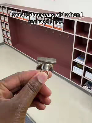 A person's hand holds a silver binder clip in front of a school locker setup. The lockers are a mix of open shelves and closed compartments, with a burgundy-colored backing visible. The text overlay reads 'I was today years old when I realized this...'.