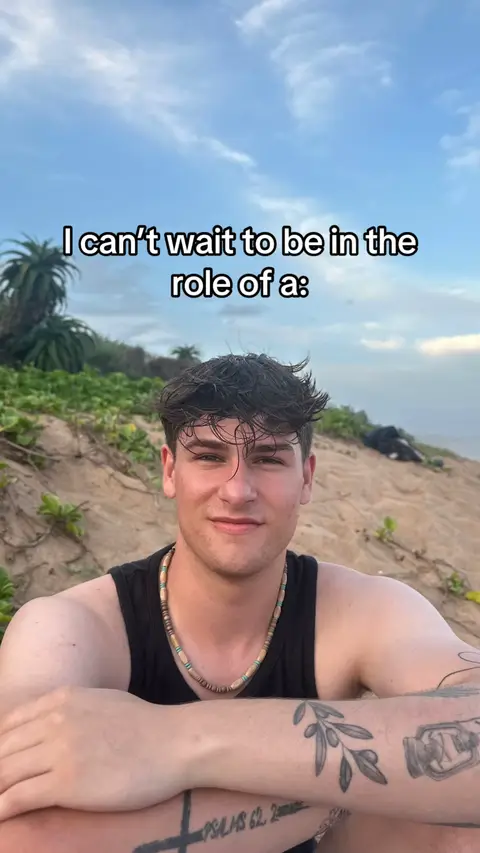 A young man with dark hair and a small tattoo on his arm is smiling while sitting on a sandy beach, with vegetation in the background and a bright sky in the background. Words are written over him in a white font.