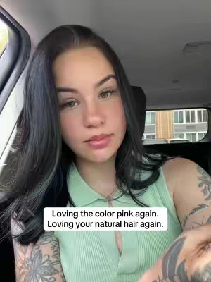A young woman with dark hair and tattoos sits in a car, looking at the camera. Text overlays read, "Loving the color pink again. Loving your natural hair again."