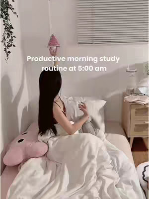 A young woman with long black hair is sitting in bed. She is wearing a simple top and the bed is covered with white sheets and a plush pillow. The room is bright and airy, with a window and blinds visible in the background. The video seems to be a study vlog.
