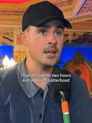 The cover image features a man wearing a black cap, speaking into a microphone. Overlayed text provides context for his discussion about Letterboxd. The background has a traditional design, possibly a cultural space, lit with warm colors. A microphone with orange and green details is prominent in the foreground.