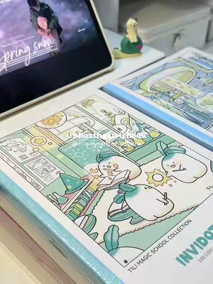 The cover image displays a close-up of notebooks on a white surface. One notebook is open, revealing a whimsical cartoon scene with characters and illustrations done in a soft color palette of blues, greens, and creams. The design of the notebook is emphasized, with the text 'TILI MAGIC SCHOOL COLLECTION INVIDOT ' and '100 GSM' visible. In the background, there is a view of a tablet displaying blurred image and a small turtle figurine, hinting at the setting.