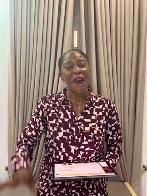 A woman is standing indoors in front of light-colored curtains. She is wearing a burgundy and white patterned dress and is holding a tablet. She is looking upward with her mouth slightly open.