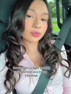 A young woman with long dark hair sits in a car, looking towards the camera. She's wearing a light pink zip-up jacket. Text overlayed on the image reads, 'How it feels to fight worldly temptations as a young Christian'. The car interior and some trees are visible in the background.