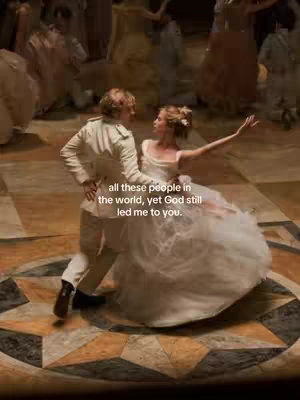 A couple dancing in a ballroom setting. The man is wearing a white jacket and tan pants, while the woman is in a white gown. They are in a close embrace, with other figures blurred in the background.