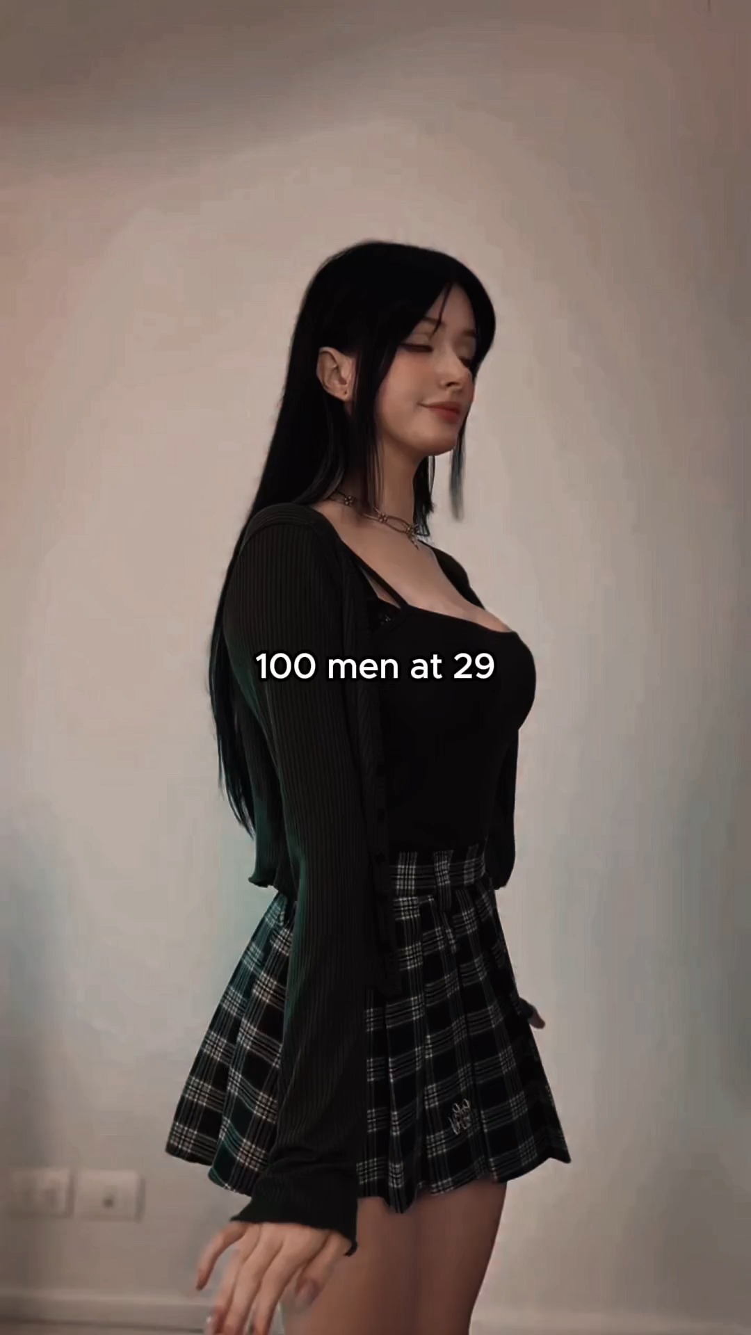A woman stands with her body angled slightly to the right, looking down with a soft expression. She is wearing a black top, a dark green cardigan, and a plaid skirt. Overlaid on the image is a white text box with the words '100 men at 29.' The background is a plain, light-colored wall.