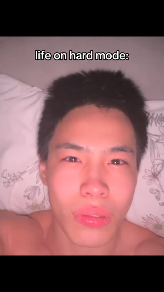 A selfie-style shot features a young man lying in bed, with the upper portion of his body visible. The man has short, dark hair, and his face is lit with a soft, slightly warm glow from the lighting. His expression appears to be a slight pout or pursed lips, looking directly at the camera. The background is blurred, with hints of a white pillow and what seems to be a patterned surface. Overlayed text reads, 'life on hard mode:' in a clean, sans-serif font.