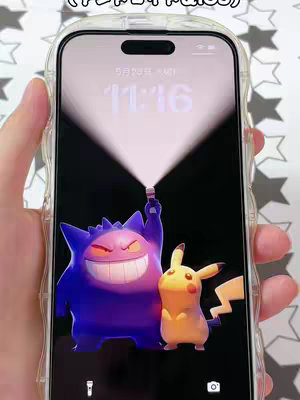 The image showcases a smartphone held by hand at a slight angle, displaying a custom live wallpaper. The lock screen shows the time and date, set against a black background. Two prominent Pokemon characters are illustrated: Gengar, in purple, grinning and holding what appears to be a glowing light, and Pikachu, in yellow, standing next to him. The phone is in a clear, protective case.