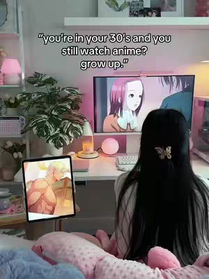 A woman with long black hair sits at a desk, facing away from the camera. She is in a well-lit room with a monitor displaying anime, a tablet showing an anime character, and other decorative items. The image is warm-toned and inviting.