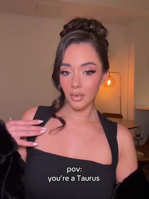 A woman with dark hair styled in a bun is centered in the frame. She is looking towards the camera. The background features a warm-toned room with a table and a lamp. Text overlay reads, "pov: you're a Taurus".