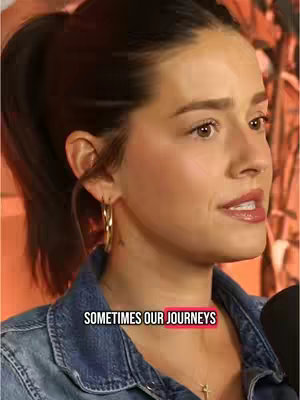 A medium shot of a woman with dark hair pulled back in a ponytail, wearing a denim jacket and gold hoop earrings. She is speaking into the camera. Text overlay 'SOMETIMES OUR JOURNEYS' in a white font with a red background.