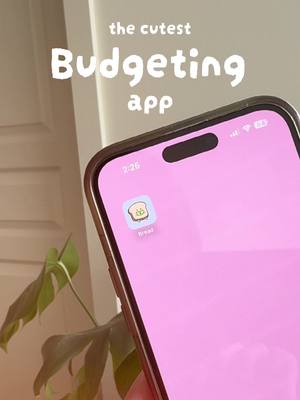 The image shows a smartphone held at an angle, displaying an app icon with a cartoon bread slice. The phone has a pink background, and text overlays that say 'the cutest' and 'Budgeting app' in a playful font. A plant is visible in the background.