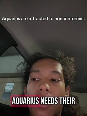 A selfie-style shot featuring a young person facing the camera, framed from the chest up, taken inside a vehicle. The person, who appears to be male, has their gaze slightly averted, off to the side. Curly hair frames their face. Text overlays are present, with the phrase "Aquarius needs their" is visible at the bottom of the screen, placed within a black rectangle with a pink outline; and "Aquarius are attracted to nonconformist" at the top of the screen. The background is indistinct, suggesting the interior of a car, with shadows and muted tones dominating.