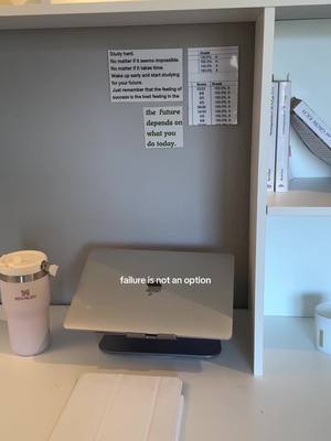 An office setting. A gray wall with motivational post-its, with a MacBook on a stand in front of it with the phrase, "failure is not an option" on it. Other decor includes a Stanley water bottle, pencil, and a couple of books.