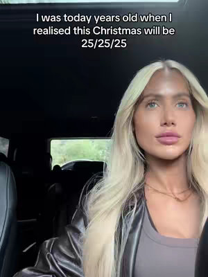 A woman with long blonde hair and light skin is seated inside a car, looking directly towards the camera. She is wearing a black leather jacket over a gray top. The background shows part of the car's interior and a blurred view of some foliage through a side window.