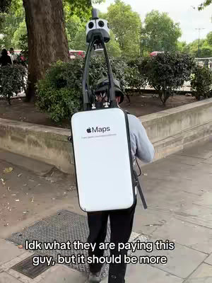 A person wearing a backpack with a large white panel that says "Maps" with the Apple logo, walking down a sidewalk. The backpack has a camera apparatus on top. Trees and bushes line the sidewalk.