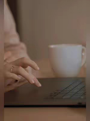 A hand with a ring on the finger is typing on a laptop, a white mug is sitting on the right side