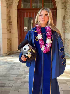 A woman in a graduation robe posing outdoors with a graduation cap and a lei. The background features a building entrance with a wooden door.
