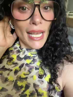 A close-up shot of a woman's face, slightly angled from the right, with focused lighting. She has large, expressive brown eyes, outlined with makeup, and is wearing large-framed glasses. Her curly, dark hair is visible on the right side of the frame. She is wearing a patterned shirt with yellow, purple, and brown details. The background is out of focus.