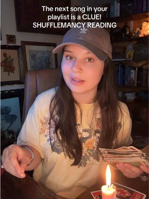 A woman with long brown hair wearing a grey baseball cap is seated at a table, holding a deck of tarot cards and gesturing with her hands. A lit candle is on the table in front of her, and in the background are bookshelves and framed pictures. The scene is warmly lit.