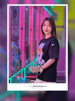 A young woman with long brown hair is standing in front of a claw machine game. The background features bright pink lights and reflections. She is wearing a black t-shirt with a cartoon graphic. The image is framed with a white border, and the text "Shot on iPhone 15 Pro Max. Apple" is visible.