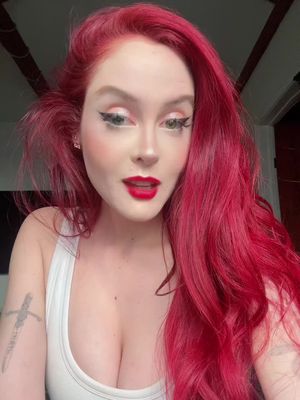 A woman with bright red hair, heavy eye makeup, and red lipstick is looking at something off-camera. She wears a white tank top and has a tattoo on her upper arm. The background is partially visible, hinting at an indoor setting.