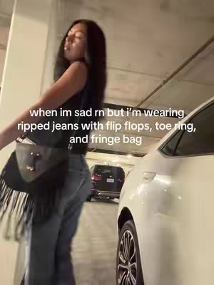 A young woman stands in a parking garage, looking slightly away from the camera. She is wearing a black top, ripped jeans, and a shoulder bag. The background includes a white car and a dark-colored SUV, suggesting a parking structure environment.